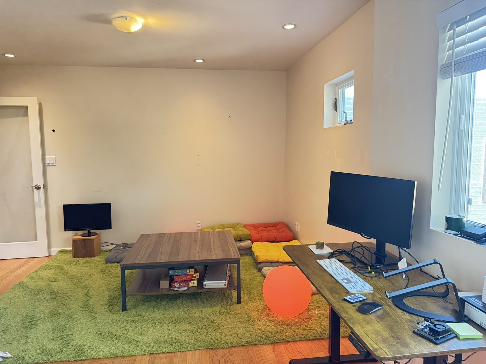 A workroom with a green shag rug, floor cushions, a low table stacked with board games and a standing desk.