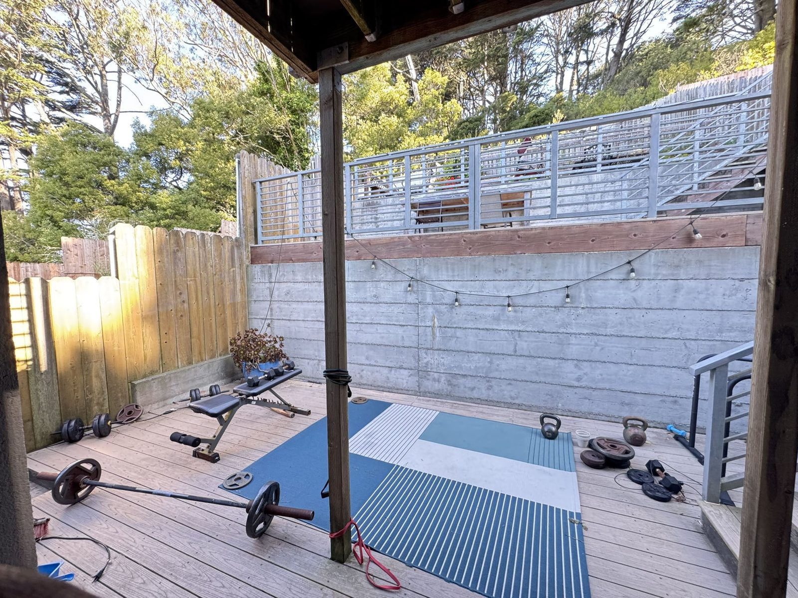 The back deck set up as an outdoor gym with a barbell, kettlebells, a bench and mats, under eucalyptus trees.